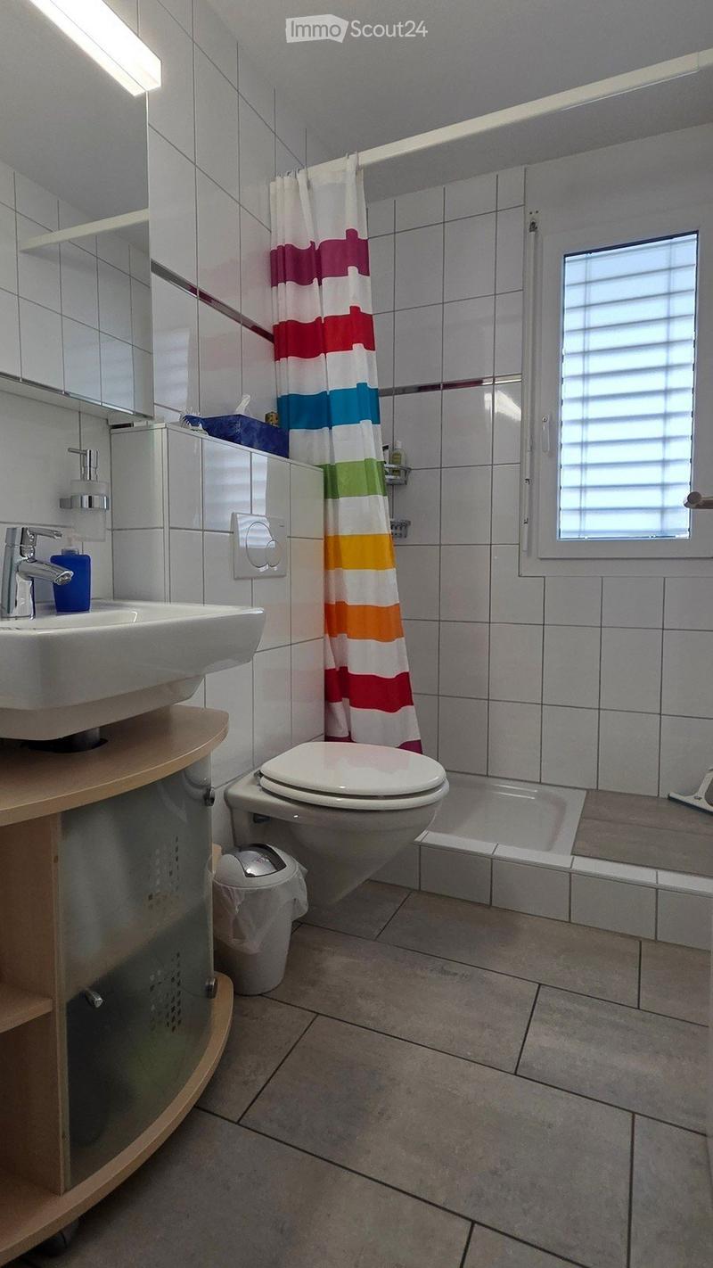 Modern bathroom with white tiles, a sink with a modern faucet, a toilet, a bathtub with a colorful shower curtain, and a small window with blinds.