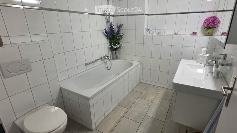 Modern bathroom with white tiles, bathtub, toilet, sink, and decorative flowers on the windowsill.