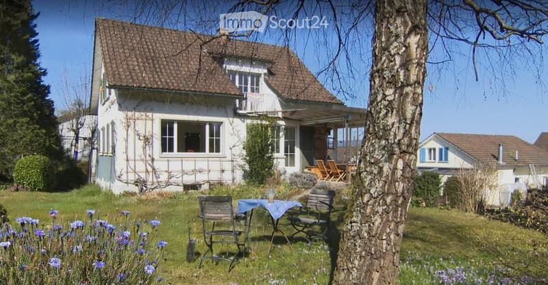 Garden with a table and chairs, surrounded by flowers and trees, with a house in the background.