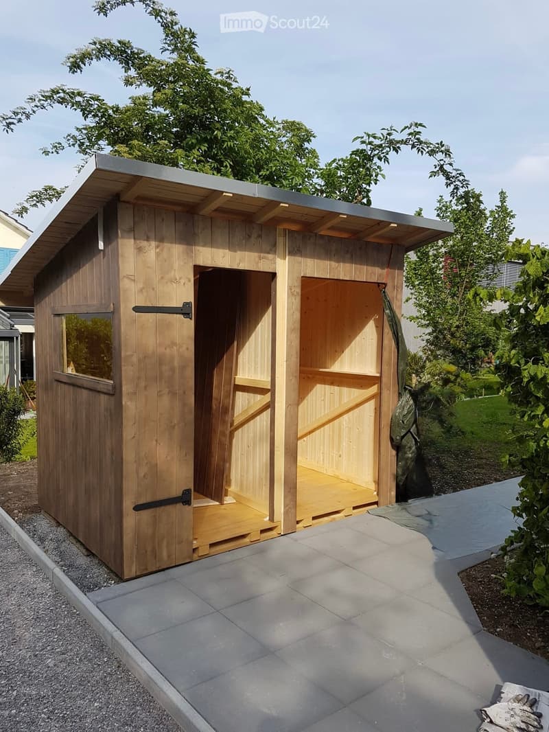 A wooden shed with a grey metal roof, open wooden doors, and a small window, situated on a paved area surrounded by greenery.