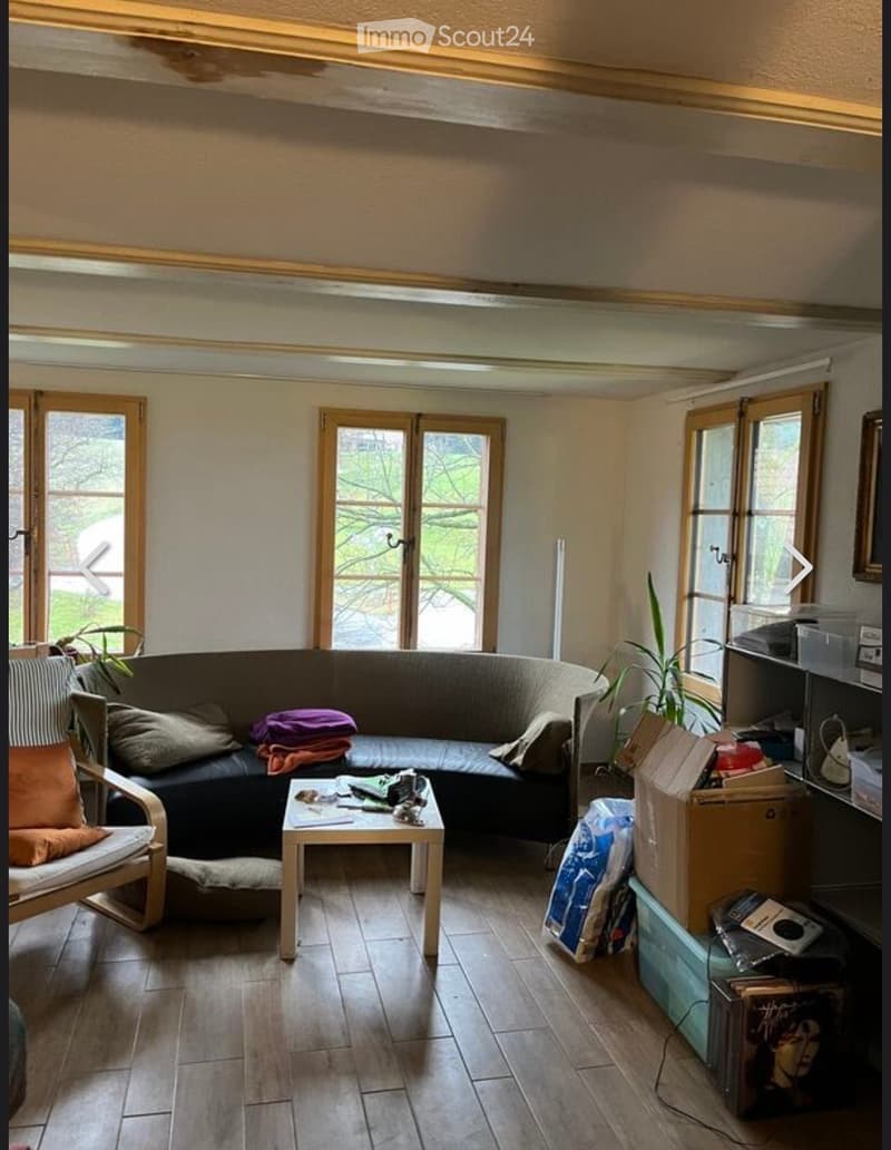 Living room with a curved sofa, coffee table, armchair, and various items including books and boxes. Large windows and wooden flooring are visible.