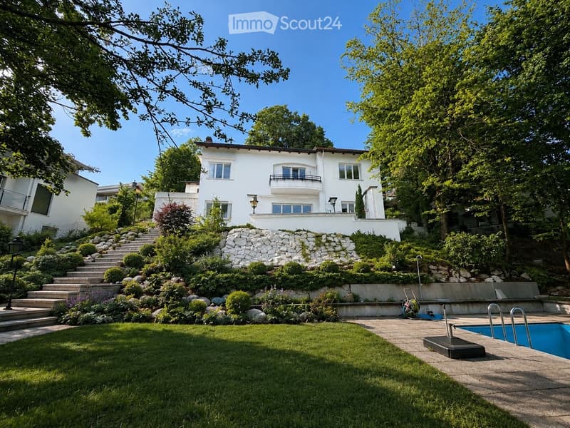 A well-maintained garden with a swimming pool, steps leading up to a white house with balconies, surrounded by lush greenery and trees.