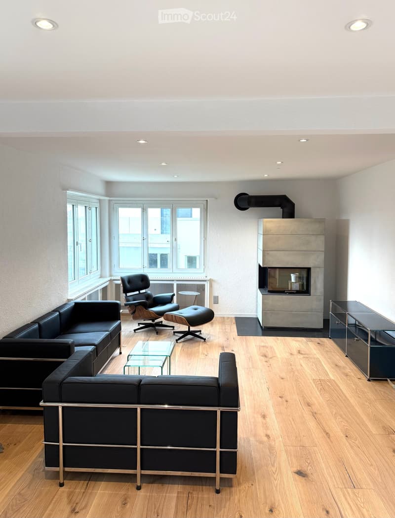 Modern living room with wooden flooring, large windows, a fireplace, a black sectional sofa, a leather chair, an ottoman, a glass coffee table, and a sleek cabinet.