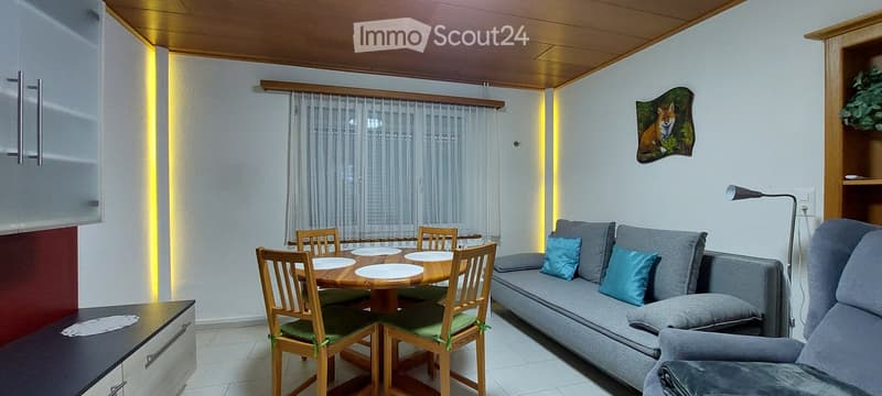 A cozy living room with a grey sofa, wooden dining table, chairs, and a floor lamp. The room features a wooden ceiling, white walls, and a decorative fox painting.