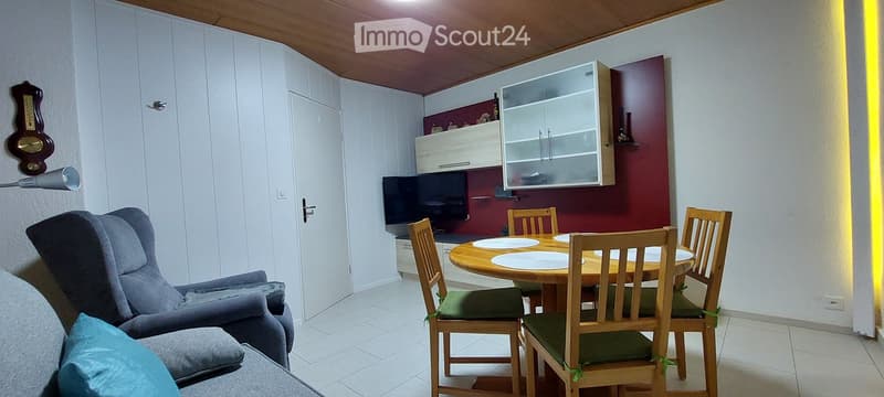 A cozy living and dining area with a grey sofa, wooden dining table with chairs, a television, and a wall-mounted cabinet.