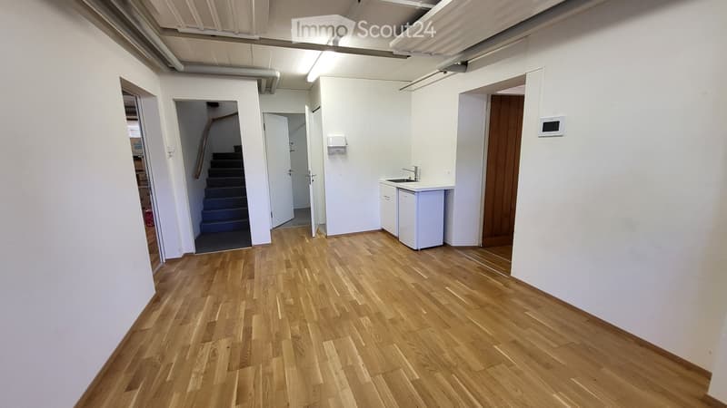 Empty room with wooden flooring, white walls, and several open doors leading to other areas, including a staircase and a small kitchenette area.