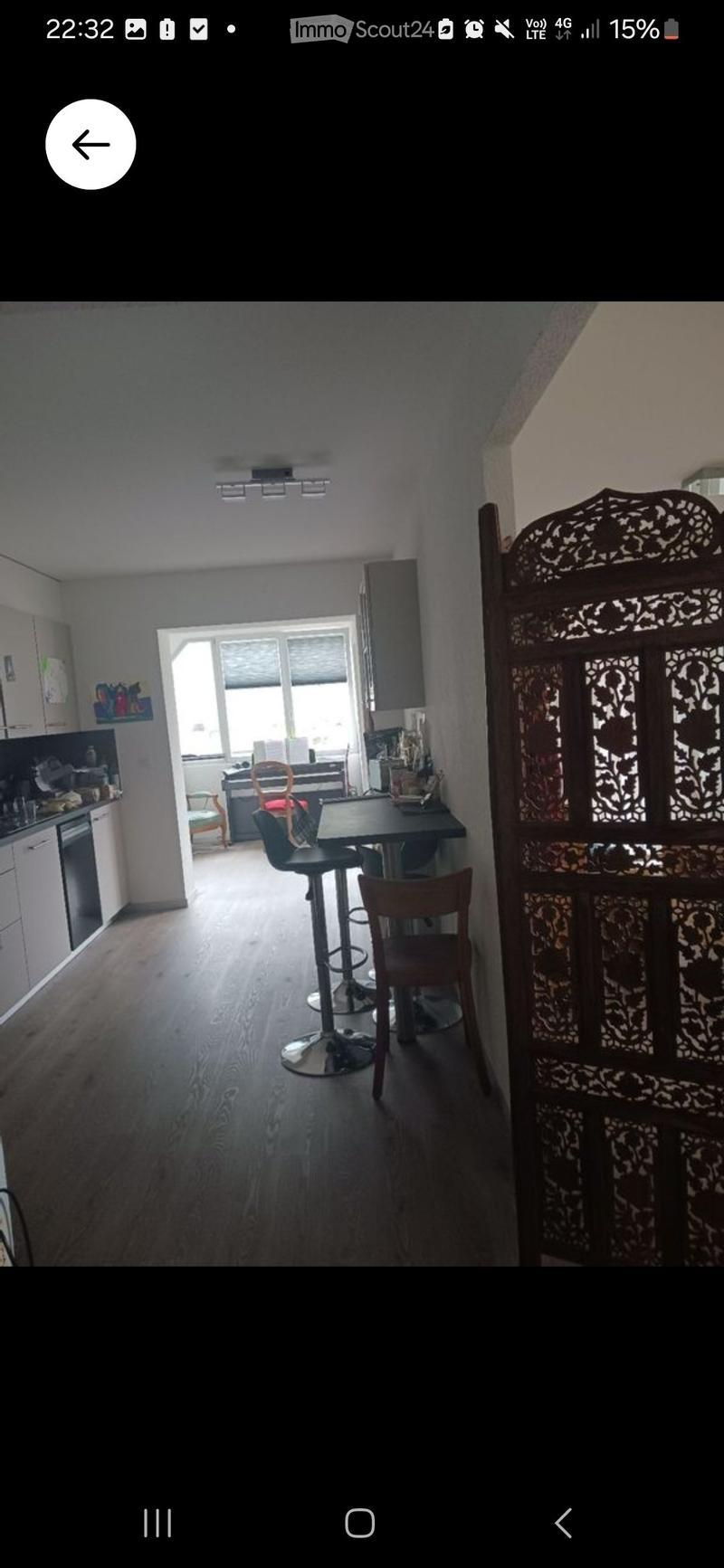 A modern, open-plan living and dining area with wooden flooring, white kitchen cabinets, a dining table with chairs, and a decorative screen. The space is well-lit with natural light from large windows.