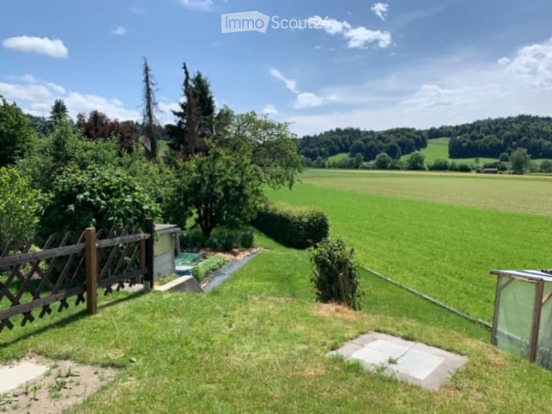 A well-maintained garden with a wooden fence, lush green grass, trees, and a small shed. The area features a clear view of a vast green field and distant hills under a bright blue sky.