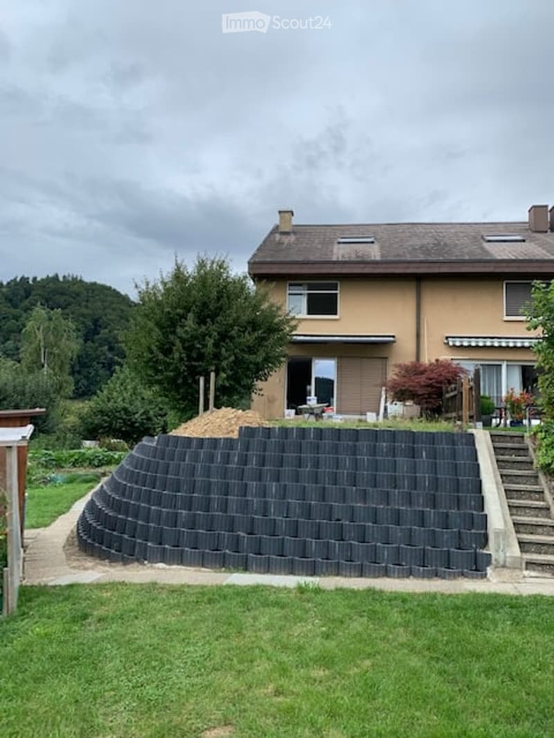 A house with a well-maintained garden, featuring a curved retaining wall made of dark bricks, a staircase leading up to the house, and a small patio area with outdoor furniture.