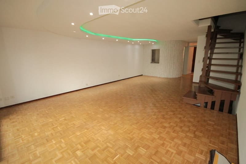 Empty room with wooden flooring, white walls, and a staircase leading upwards. The ceiling features integrated LED lighting.