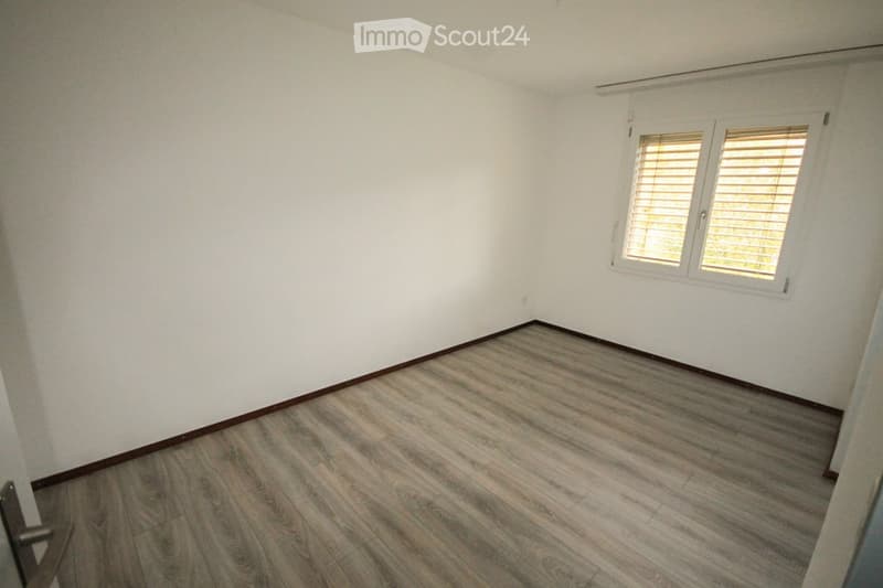 Empty room with light-colored wooden flooring and white walls, featuring a window with blinds on the right side.
