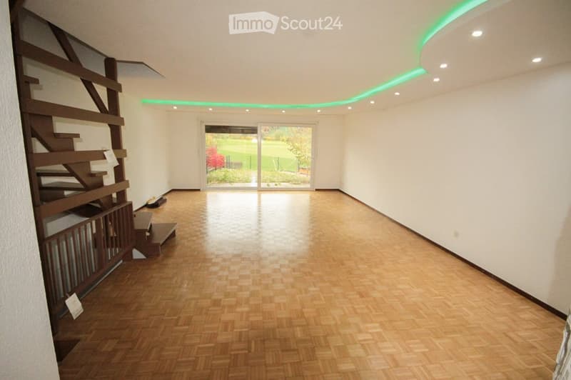 Empty room with wooden flooring, white walls, and a staircase leading upwards. A large window with a view of a green outdoor area is present.