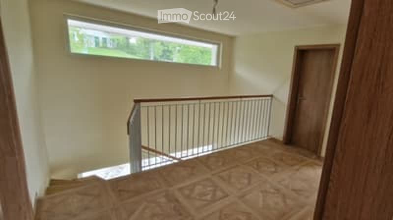 Empty hallway with wooden flooring, a metal railing leading to an upper level, and a large window letting in natural light.