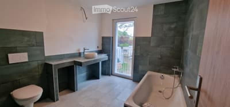 Modern bathroom with dark green tiles, a bathtub, toilet, sink, and a sliding glass door leading to a balcony.