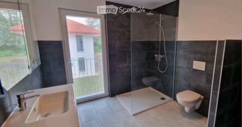 Modern bathroom with dark grey tiles, a large window with a view of a balcony, a white sink with a chrome faucet, a toilet, and a glass-enclosed shower stall with a modern showerhead.