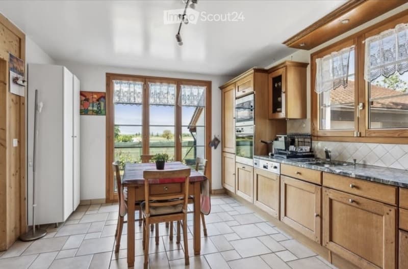 Bright kitchen with wooden cabinets, stainless steel appliances, and a dining area with a table and chairs. Large windows provide natural light and a view of the outdoors.
