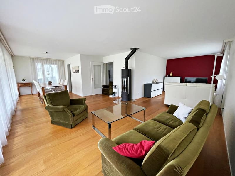 A spacious living room with a green sofa, matching armchair, glass coffee table, dining area with a table and chairs, a fireplace, and a TV screen on a stand.