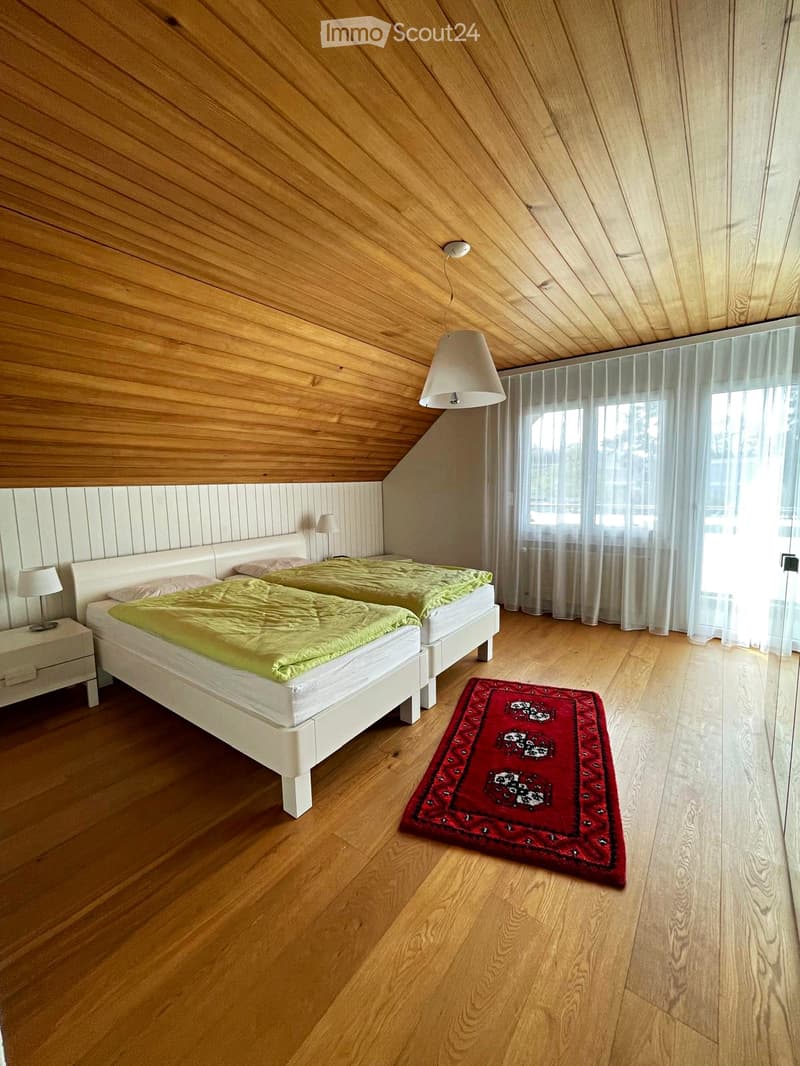 Bright bedroom with two single beds, green bedding, white nightstands, lamps, and a red patterned rug on wooden flooring.