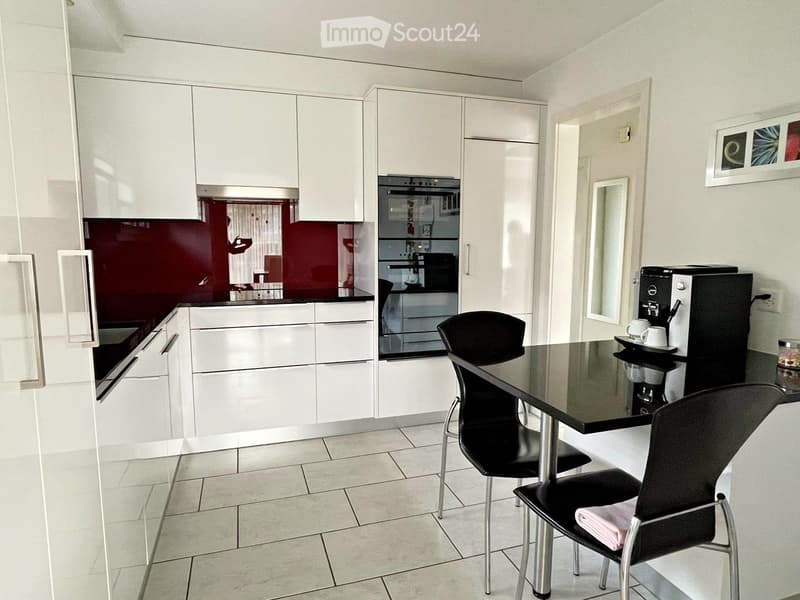 Modern kitchen with white cabinets, a black countertop, a coffee machine, a dining table with two chairs, and an oven.