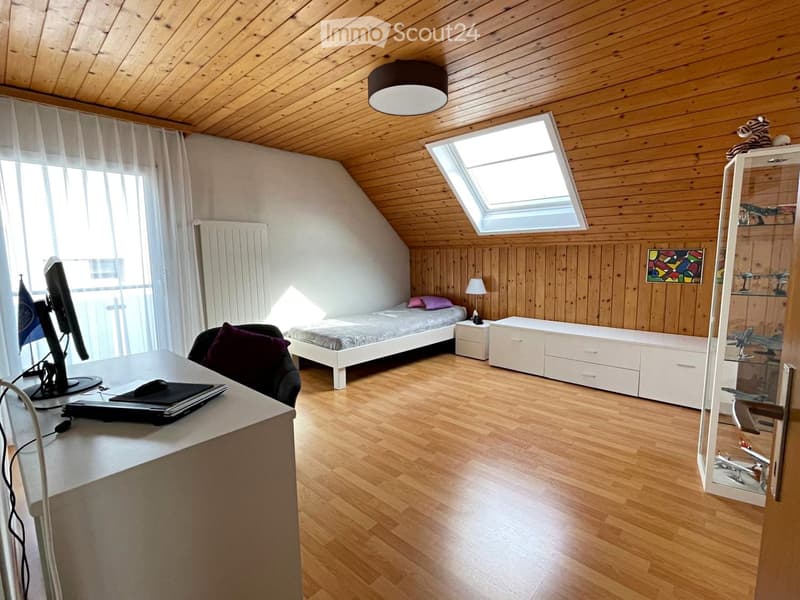 Bright bedroom with wooden walls and ceiling, featuring a bed, desk with laptop, chair, nightstand, and cabinet.