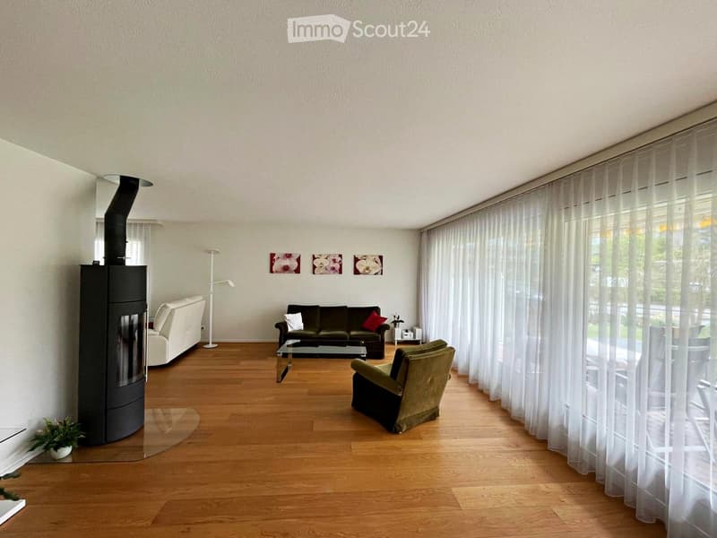 Modern living room with wooden flooring, fireplace, white curtains, and large windows. Features a cream sofa, green armchair, coffee table, and floor lamp.