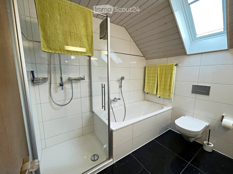 Modern bathroom with a bathtub, toilet, glass-enclosed shower, and towels hanging on the wall. The room features white tiles and a skylight window.