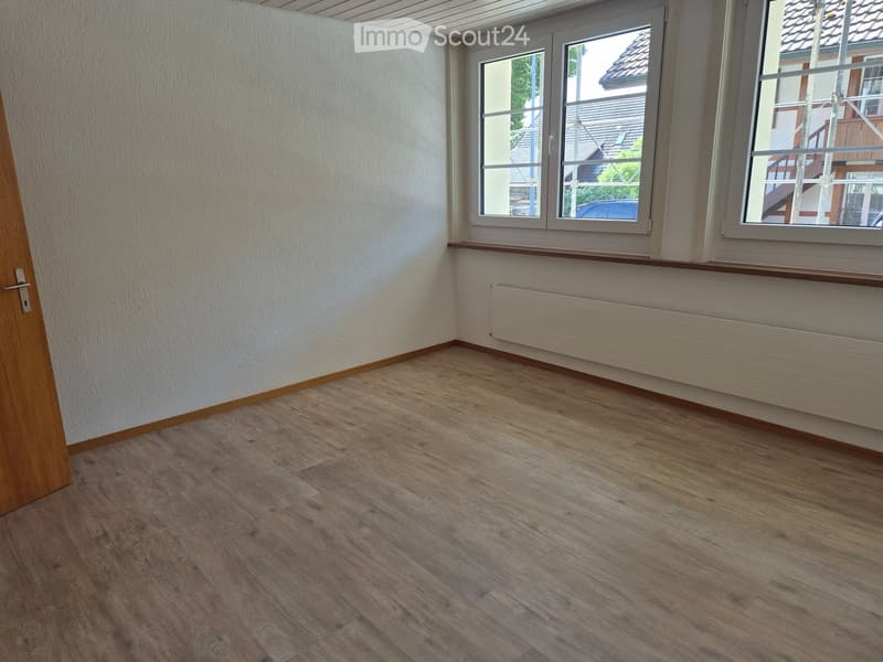 Empty room with wooden flooring, white walls, and large windows with a view of neighboring houses.