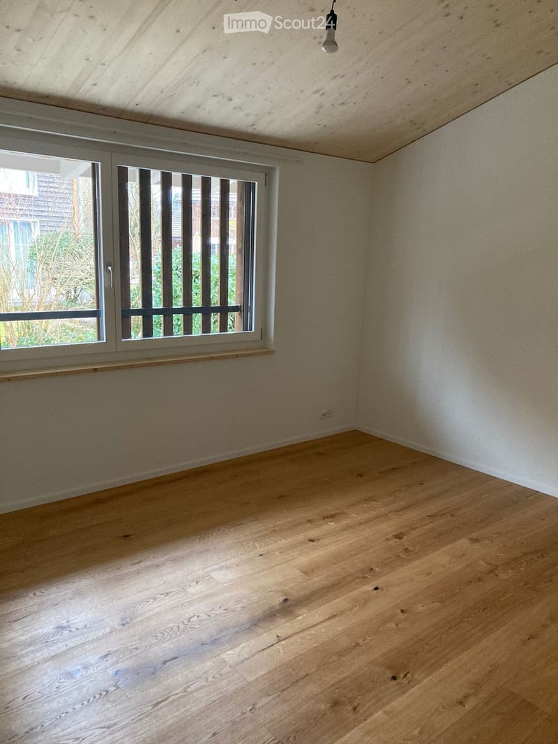 Empty room with wooden flooring, white walls, and a large window with blinds. The room has a simple, clean design with a single light fixture hanging from the ceiling.