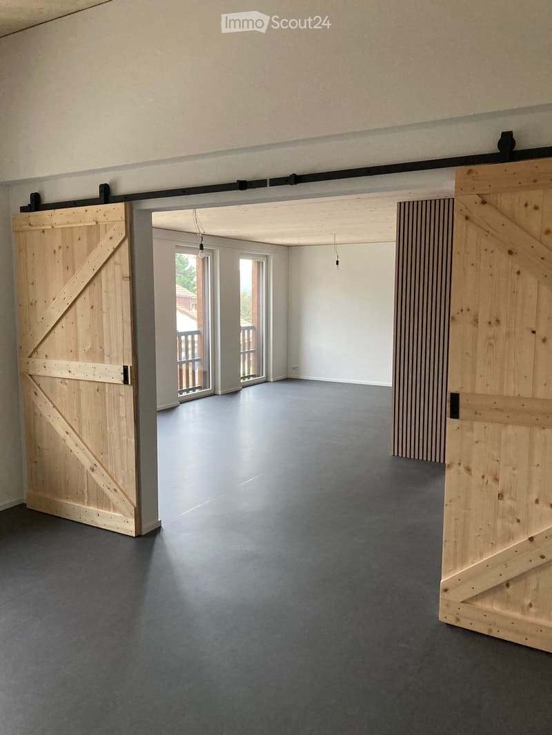 Empty room with grey concrete floor, white walls, and large wooden barn-style doors on either side, leading to an outdoor area with a wooden railing visible through the doorway.