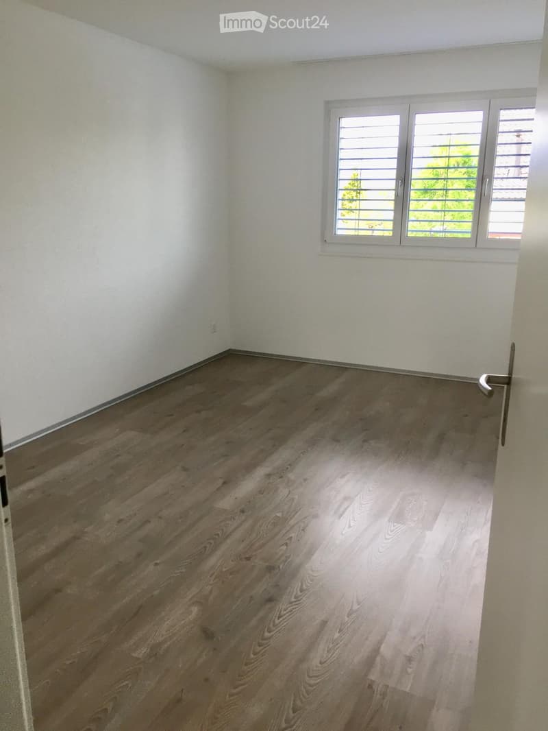 Empty room with light wooden flooring and white walls, featuring a large window with blinds and a door handle visible.