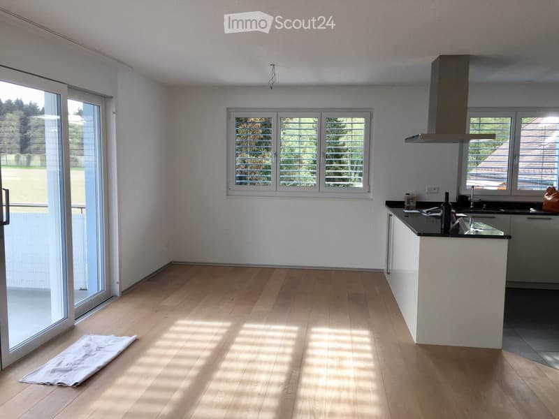 Empty kitchen with wooden flooring, white walls, large windows, and a sliding glass door leading to a balcony.