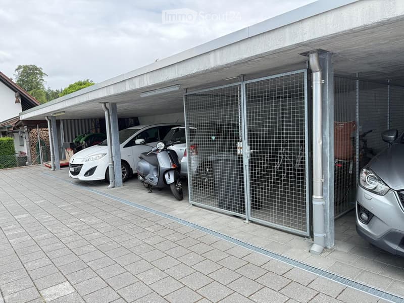 Carport with a covered area, containing a white car, a scooter, and a motorcycle, with a metal gate and a paved floor.