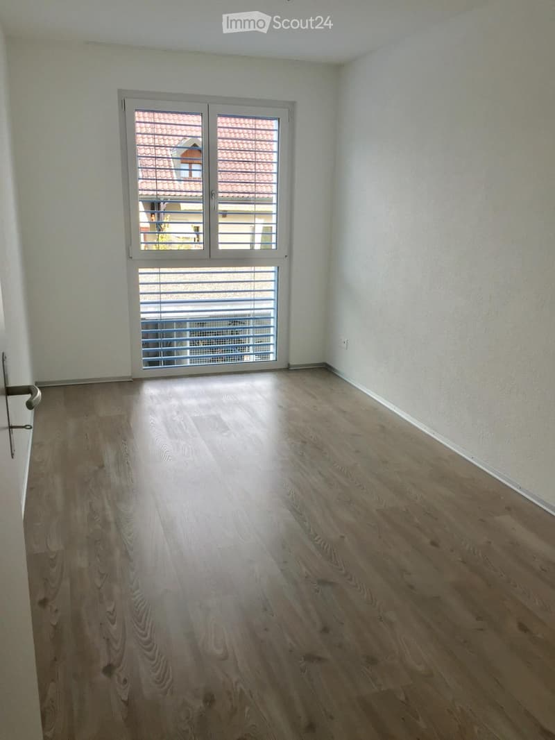 Empty room with light wooden flooring, white walls, and a large window with blinds offering a view of a tiled roof and adjacent building.