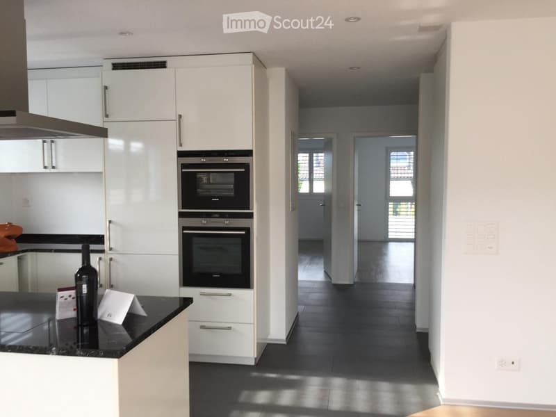 Modern kitchen with white cabinets, black countertops, and built-in ovens. A hallway leads to another room with large windows.