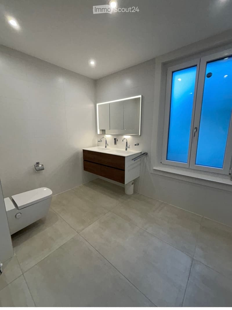 Modern bathroom with a double sink, toilet, large mirror, and built-in cabinetry. The space features tiled floors and walls with recessed lighting.