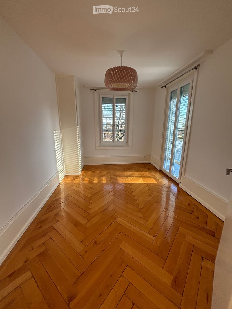 Empty room with wooden herringbone flooring, white walls, large windows, and a hanging light fixture.