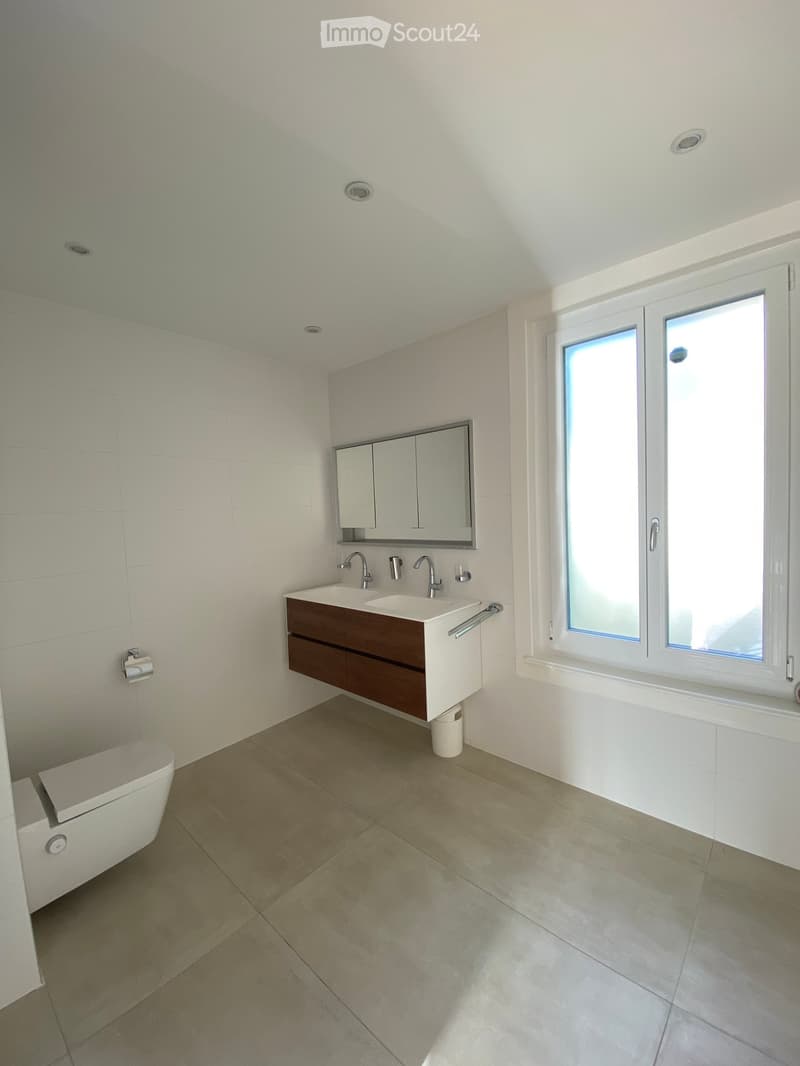 Modern bathroom with a double sink, mirror, and toilet. Light-colored tiles and a large window provide natural light.