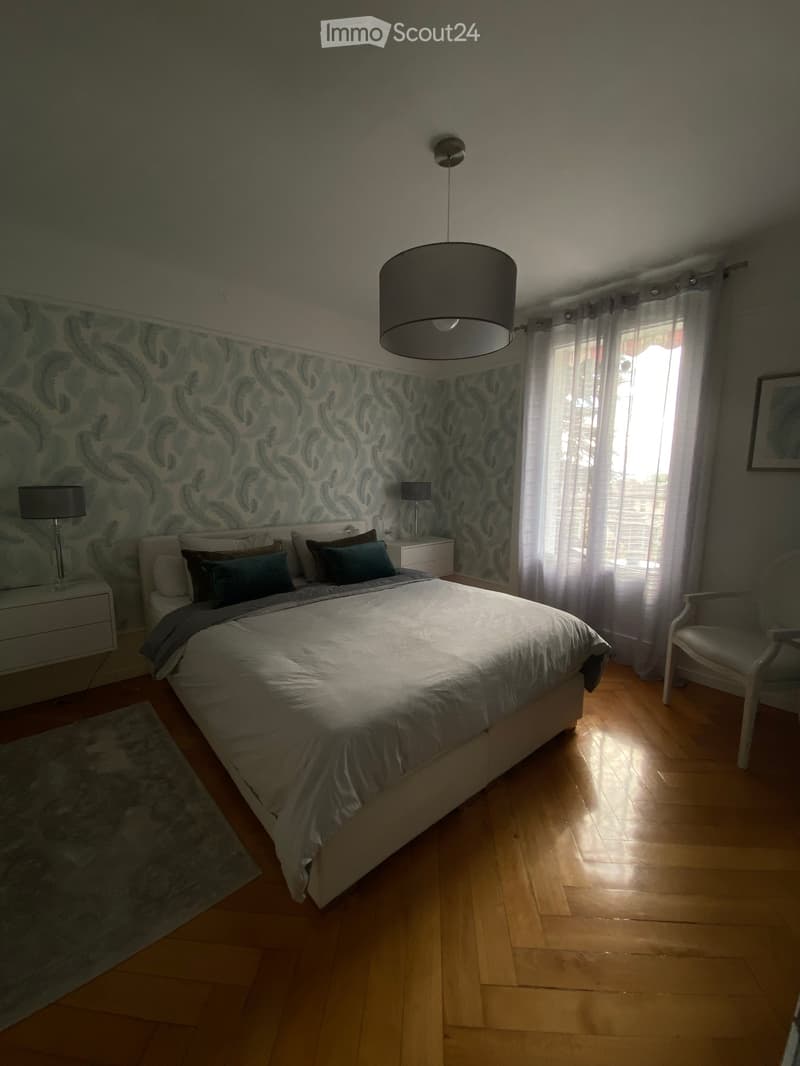 A well-lit bedroom with a large bed, nightstands, a chair, and a hanging lamp. The room features wooden flooring and patterned wallpaper on one wall.