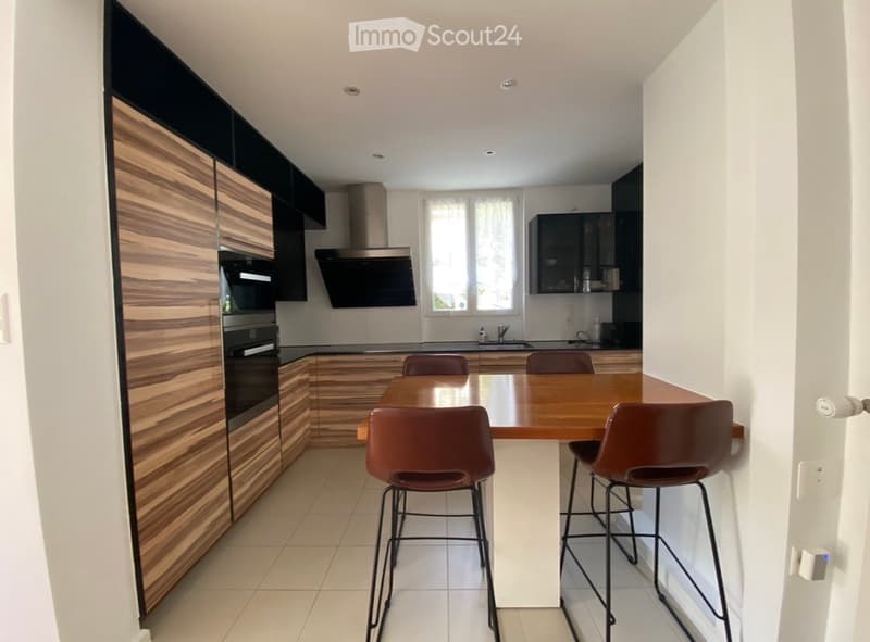 Modern kitchen with wooden cabinets, stainless steel appliances, a central island with a wooden table and four brown chairs, and a window with white blinds.