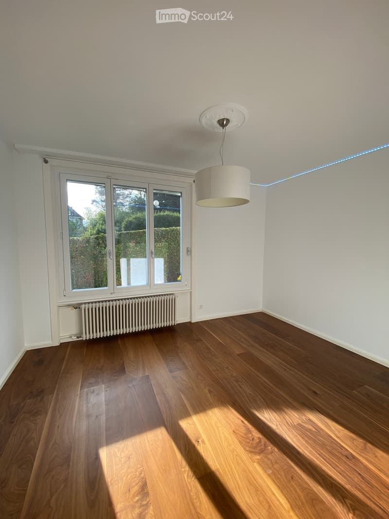Empty room with wooden flooring, white walls, large windows, and a hanging light fixture.