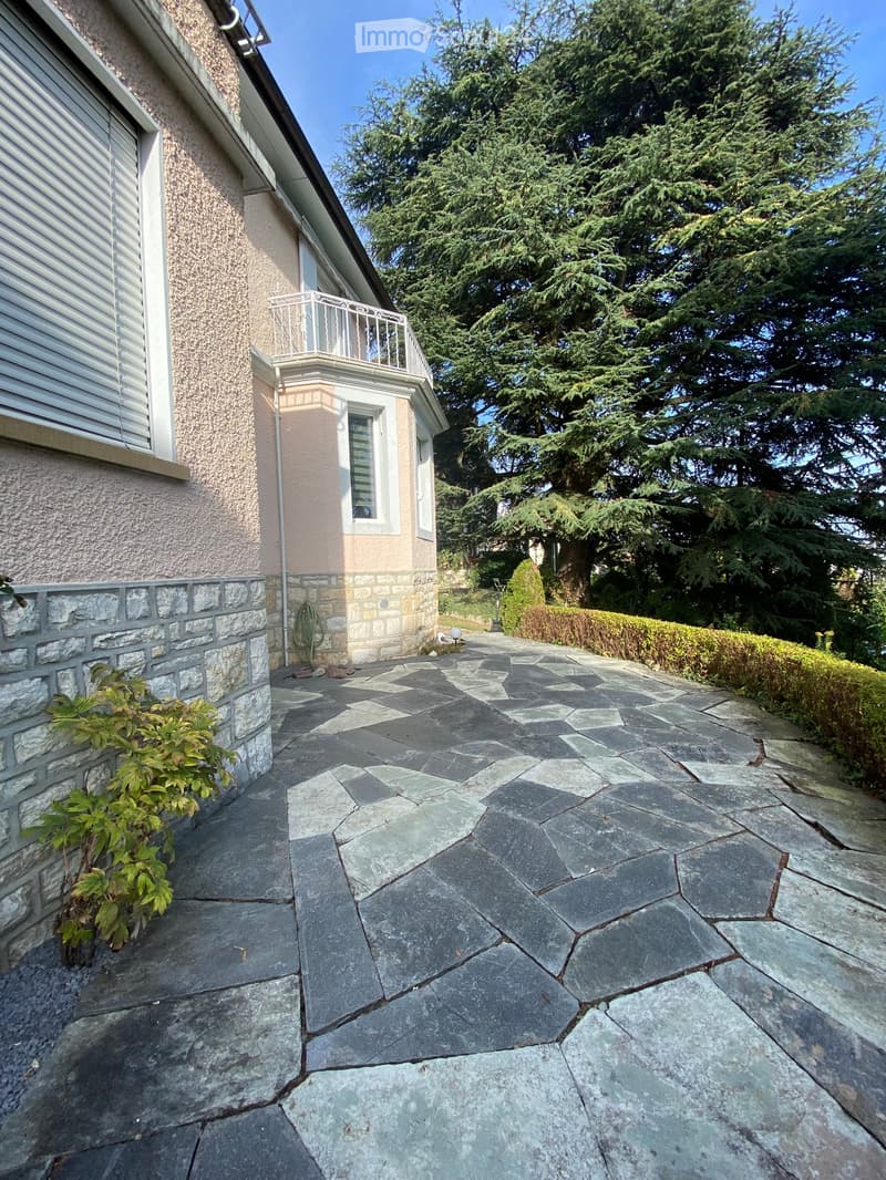 Stone-paved patio area adjacent to a house with a small balcony, surrounded by greenery and trees.
