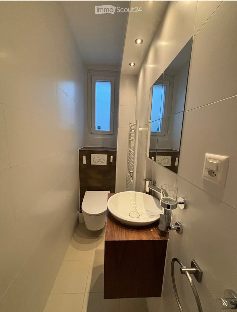 Modern bathroom with a white sink, dark wooden cabinet, toilet, and a large mirror above the sink. The space is illuminated by recessed ceiling lights and has a small window providing natural light.