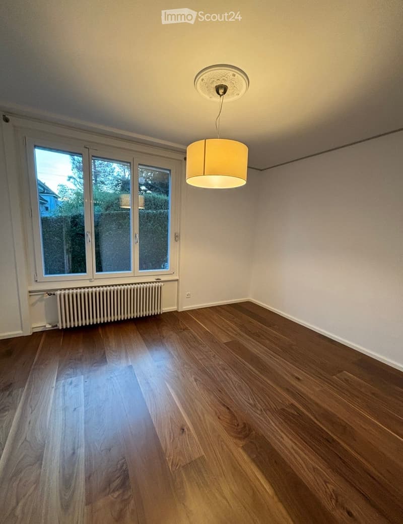 Empty room with wooden flooring, large windows, radiator, and a hanging light fixture.