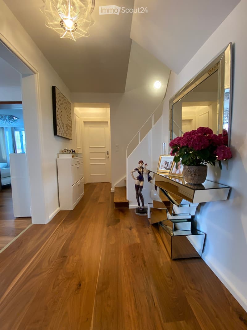 Elegant hallway with wooden flooring, modern lighting, a staircase, and decorative items including a mirror, vase with flowers, and framed photos on a console table.