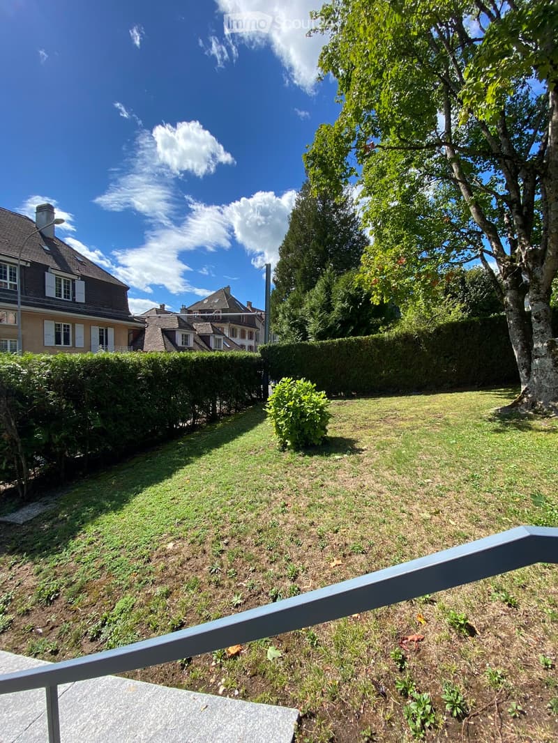 A well-maintained garden with a sloping lawn, trimmed hedges, and a large tree, viewed from a balcony with a metal railing.