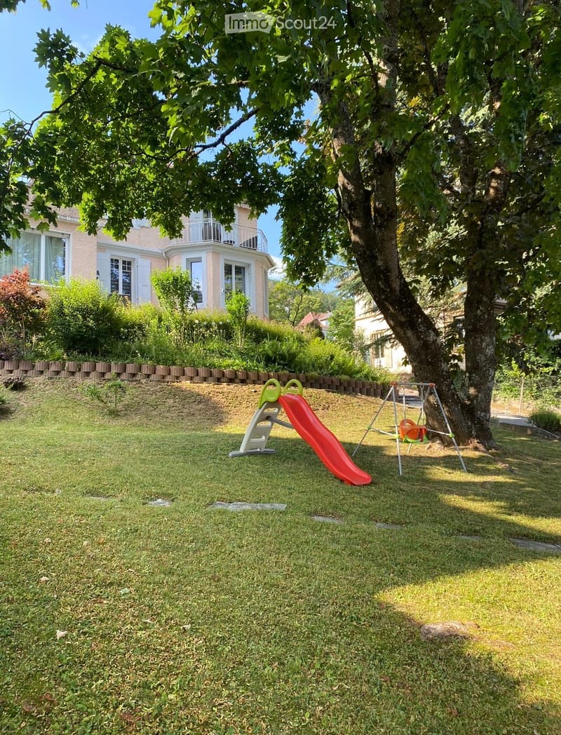 A well-maintained garden with a slide, swing, and a large tree providing shade. The lawn is surrounded by a low brick wall and a house with a balcony is visible in the background.