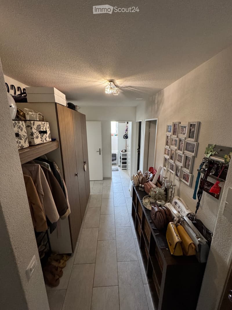 A narrow hallway with tiled flooring, beige walls, and a ceiling light fixture. A wooden wardrobe with coats and boxes is on the left, and a small table with handbags and decorative items is on the right. Several framed photos are displayed on the wall.