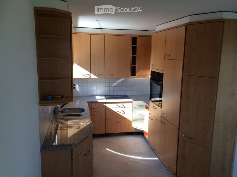 Modern kitchen with light wooden cabinets, granite countertop, sink, and built-in oven. Sunlight streams in from a window, illuminating the space.
