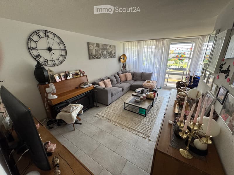 A modern living room with a grey sectional sofa, coffee table, side table, and decorative items including candles and picture frames. The room features large windows with sheer curtains and a view of the outside.