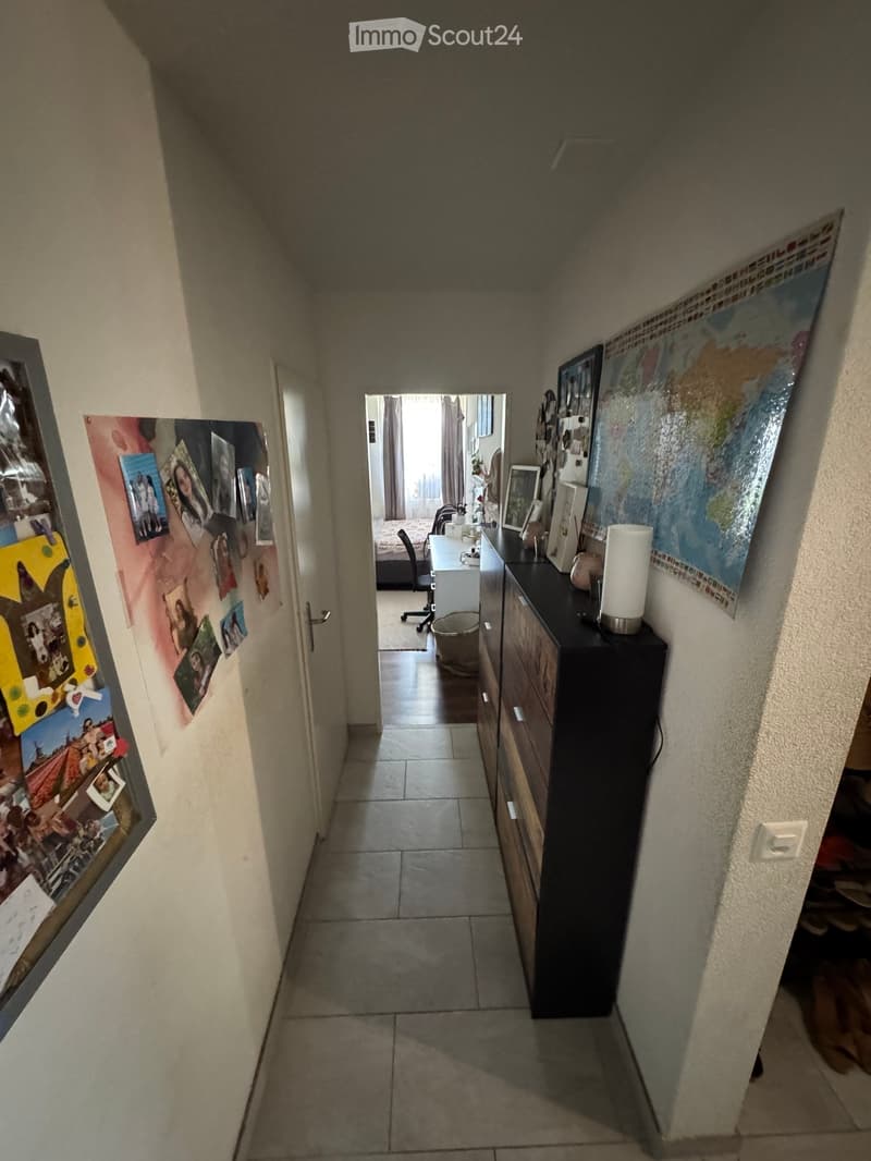 A narrow hallway with tiled flooring, light-colored walls, and a small open doorway leading to another room. The hallway features a small cabinet with drawers and a lamp on top, along with a map and framed pictures on the walls.